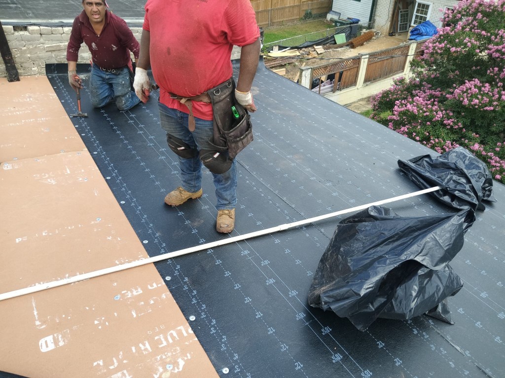 Two workers installing a rubber roof, standing on the roof surface with tools and materials, surrounded by residential buildings and greenery.